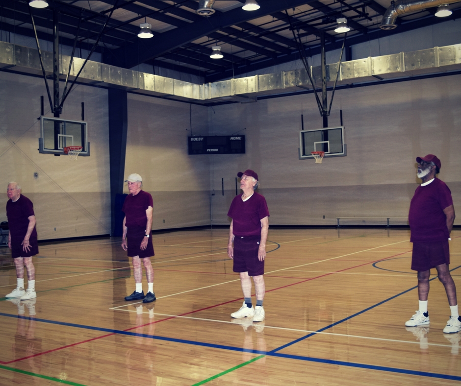 Four senior men lined up doing exercises in a gym.