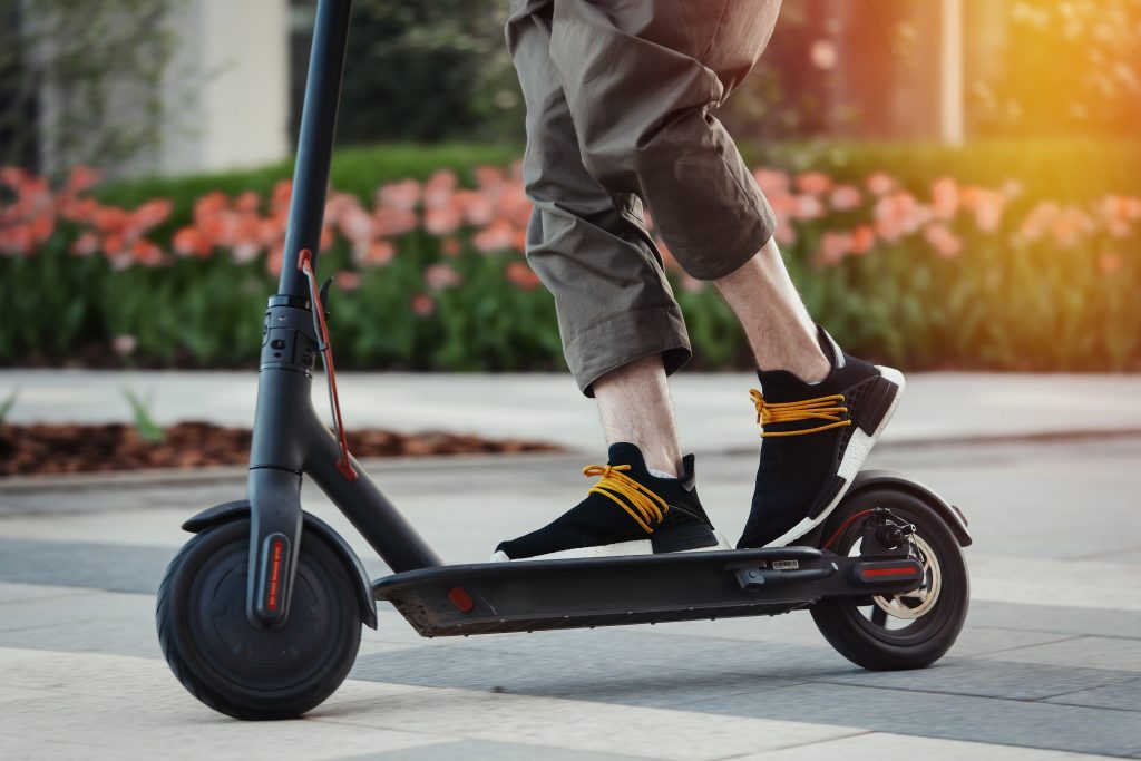 lose up of man riding black electric kick scooter at beautiful park landscape. Man is on foreground, modern building and park is on background.