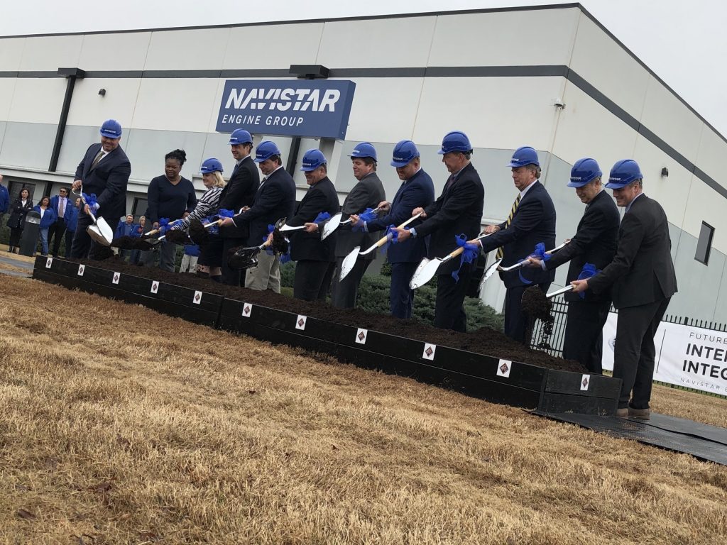 men and women wearing construction hard hats line up with shovels to scoop dirt in a ceremonial groundbreaking for Navistar