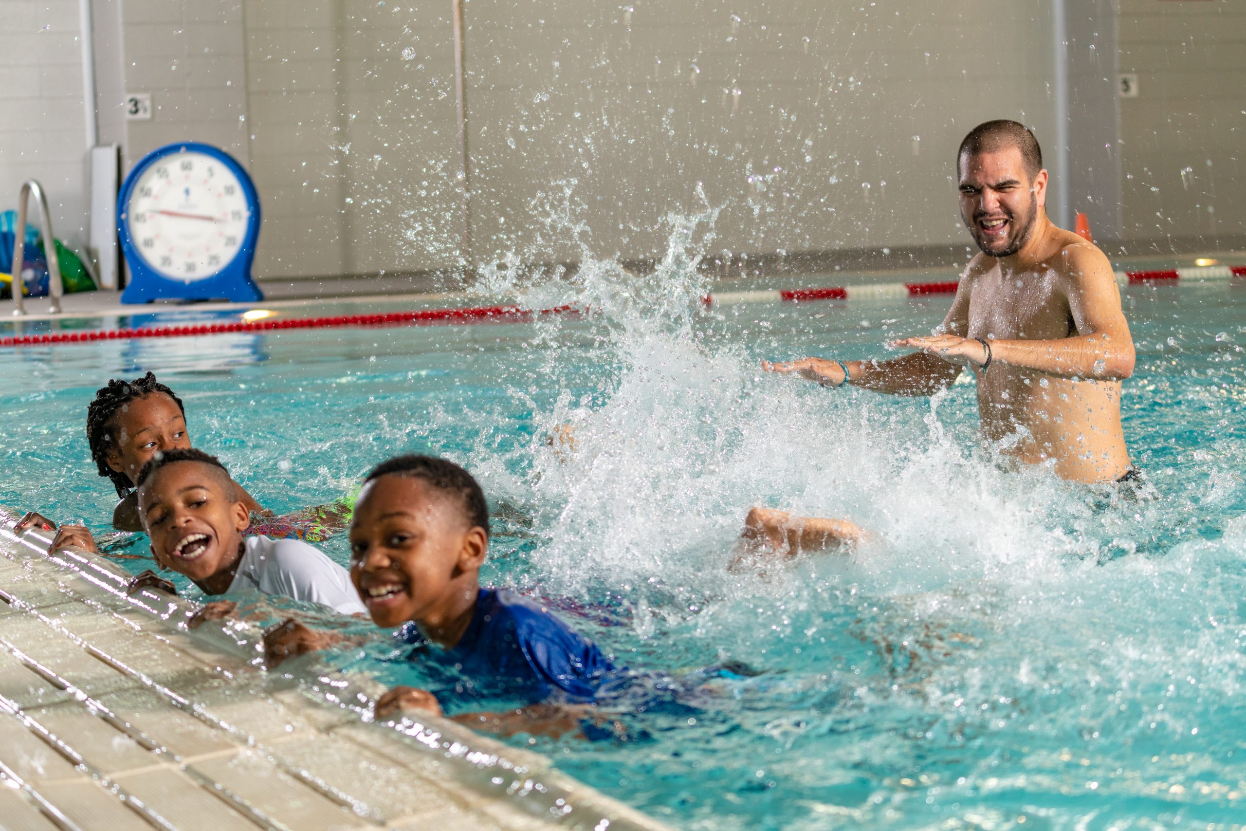 Children kicking feet from the side of the pool while instructor smiles and splashes.