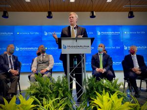 Huntsville Mayor Tommy Battle stands behind a podium and gestures while making comments at a groundbreaking ceremony for HudsonAlpha. There are people sitting in chairs behind him wearing masks and looking up at him as he speaks. There are plants in the foreground and several blue screens in the background.