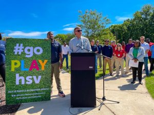 Mayor Battle at the podium in a City park with a poster announcing Go Play H-S-V for Parks & Recreation Month in July