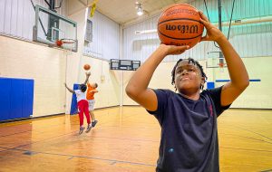 Young boy shooting a basketball inside a gymnasium. Two young boys are in the background also playing basketball on another goal.