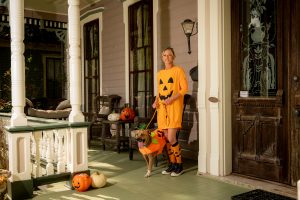 A dog and a woman are seen on the front porch of a historic home. There are pumpkins around.