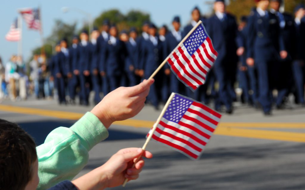 People wave flags at a Veterans Day parade. There is a group of cadets marching in the background.