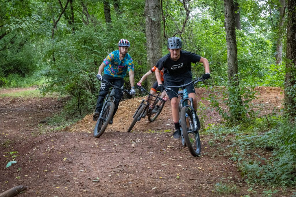 three young boys are pictured riding their bikes down a wooded path in the John Hunt Park Mountain Biking Course
