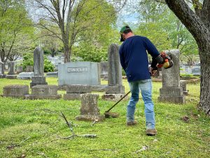 A man in a dark blue shirt, cap and blue jeans operates a weed wacker around a headstone in a city cemetery. There are various sticks and limbs laying on the ground. The grass is green and there are trees in the background.