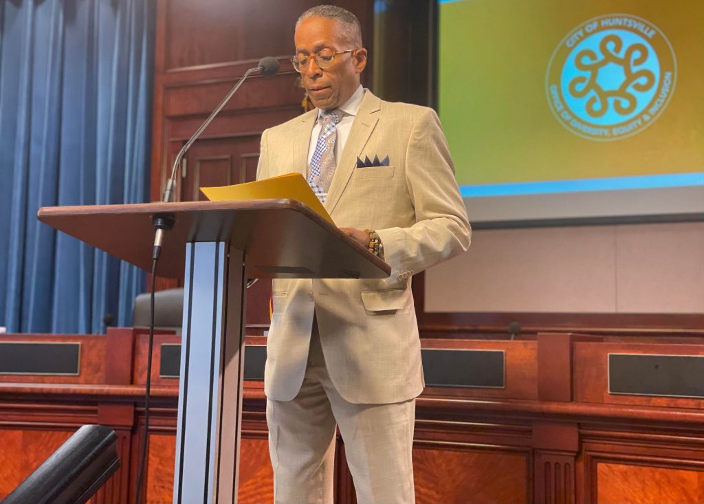 man in tan suit and glasses speaks at podium during news conference in chambers