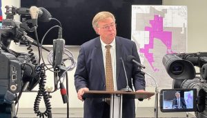 Man in suit and glasses speaks at podium with microphone during news conference with cameras