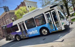 An Orbit bus is seen on a street with a tree and brick building in the background. The bus is blue, purple and white and has "orbit" on the side.