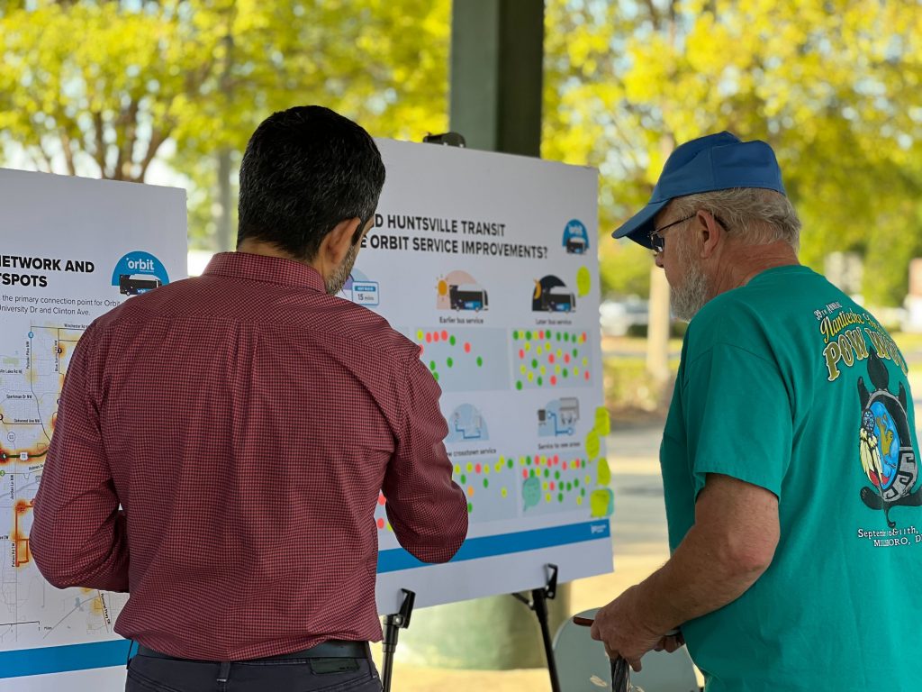 Two men look at a board asking for feedback on transit improvements. Both men have their backs turned. One is wearing a red shirt. The other a green shirt and a blue cap.