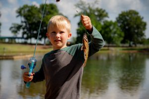 A small child holds up a fish caught during a fishing rodeo at Brahan Spring Park. He's holding a fish in one hand and rod in another.
