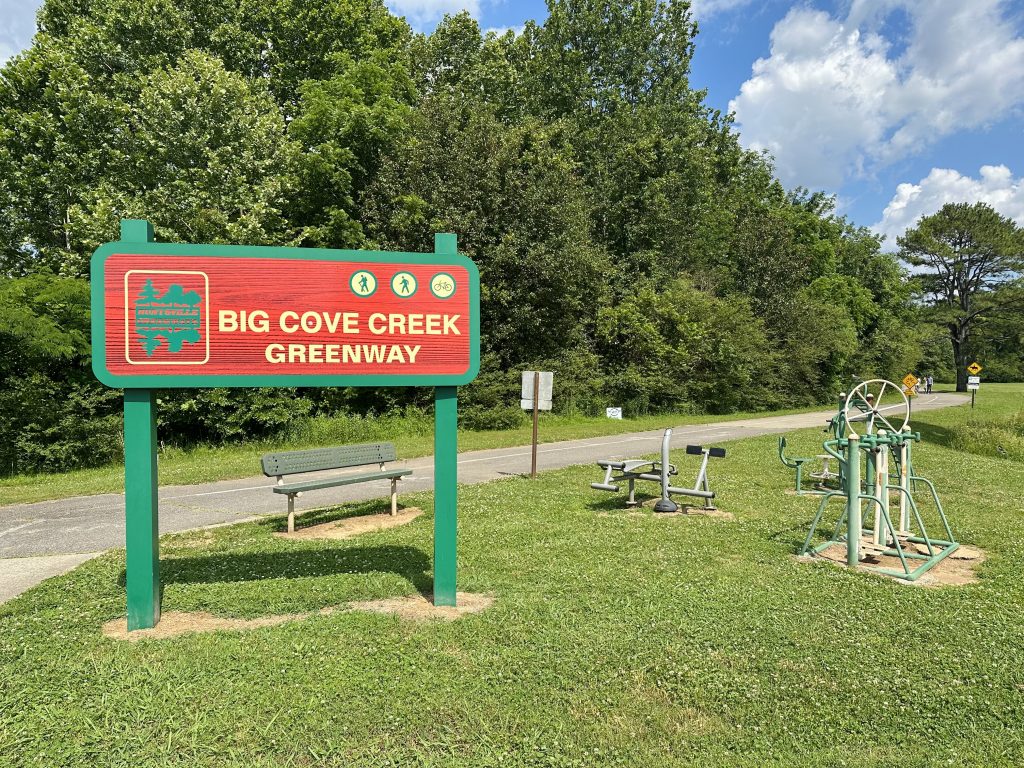A photo of the trail head of Big Cove Creek Greenway. There is a red and green sign with the name of the greenway, a bench and bike rack visible in the photo. There is also green grass and trees in the background.