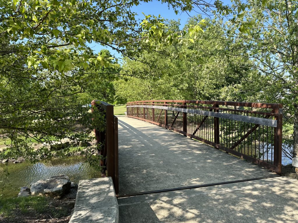 A photo from the Gateway Greenway, which includes a bridge going over a creek with a brown handrail. There are trees all around and a creek under the bridge.