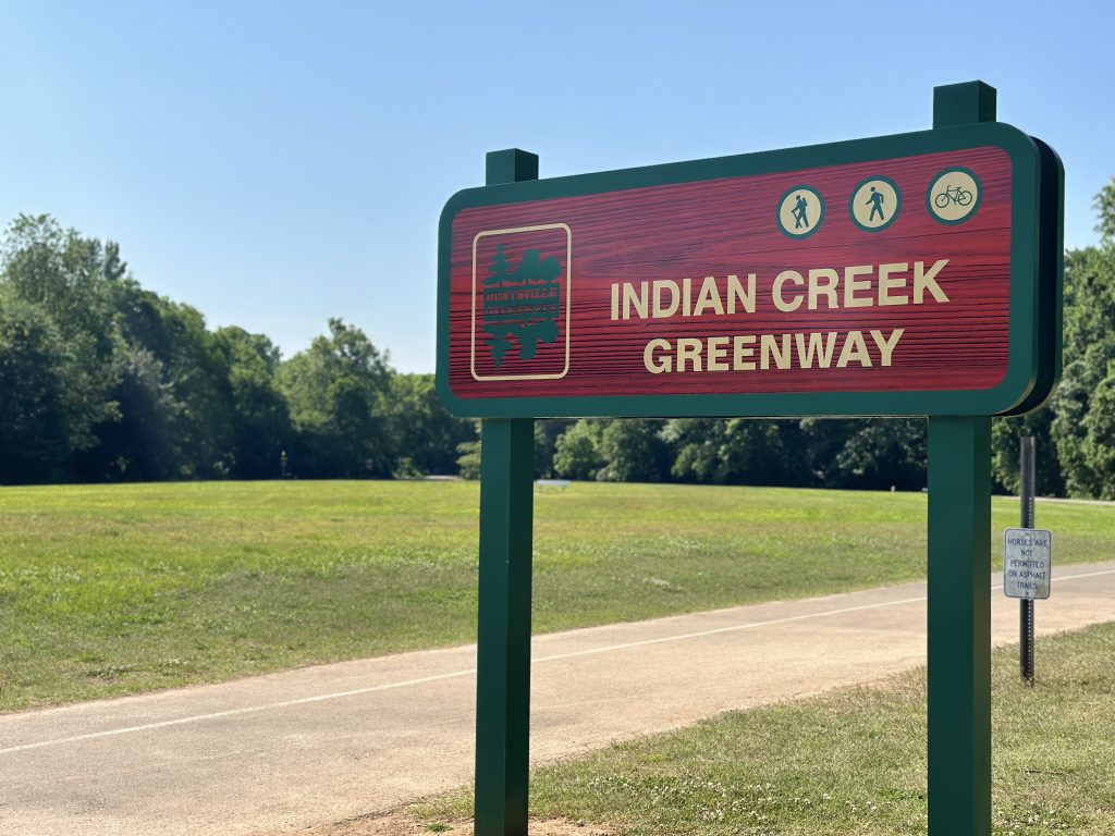 A sign at the trail head of the Indian Creek Greenway in Huntsville. The sign is red and green. There are trees and grass in the background and the sky is clear.