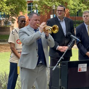 A man in a gray suit plays a golden trumpet at a podium as three people stand behind.