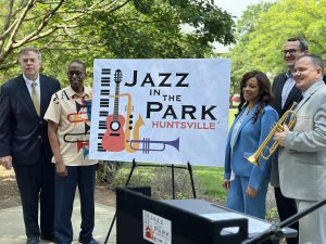 A group of five people stand next to a sign that says Jazz in the Park sign in Big Spring Park.