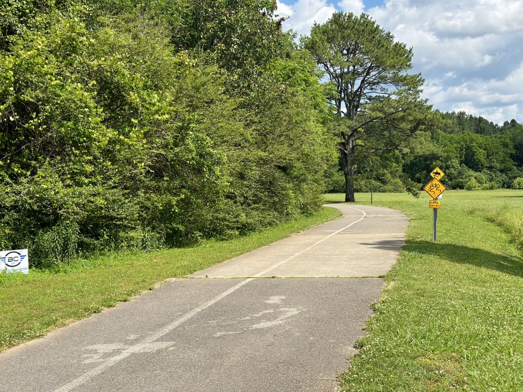 A paved greenway is seen next to a grove of trees. To the right is a yellow sign with a bicycle and another with a man on horseback. There is green grass to the right of the pathway.