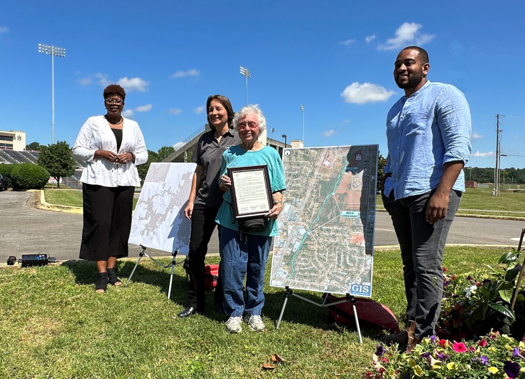 three women and one man standing outdoors with posters showing the route of the new Meek Greenway