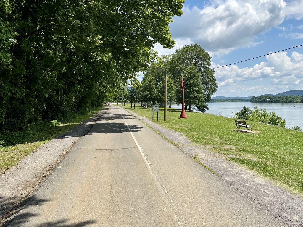 A picture of the Tennessee River Greenway. There is paved trail with the river to the right. There is also a bench facing the river and many trees to the left of the path.