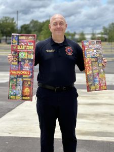 A man with a bald head and blue shirt holds up a box of fireworks in each hand.