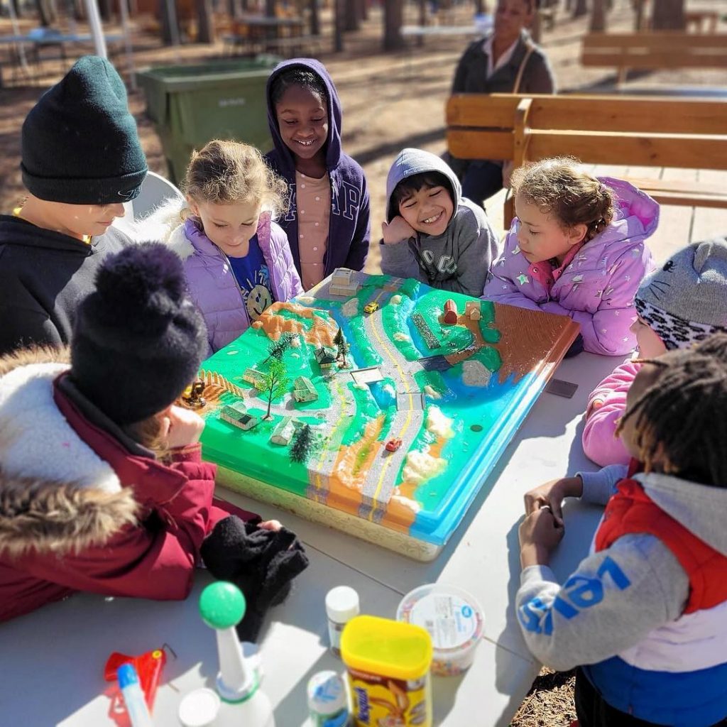 Children sitting outdoors gather around a table and play a board game showing the topography of land, rivers, mountains