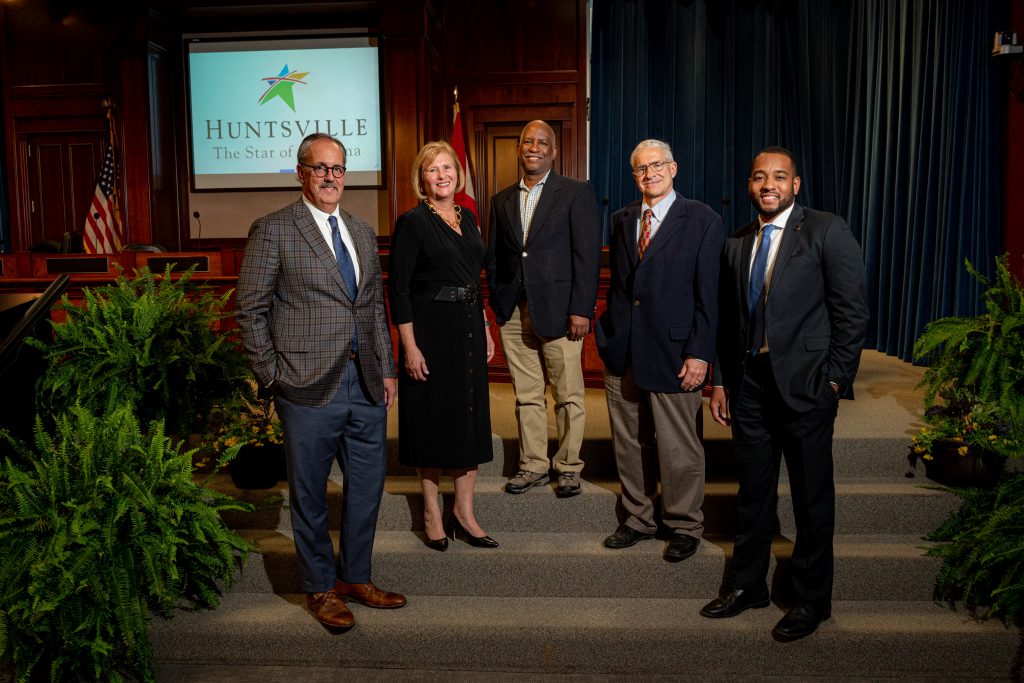 four men and one woman stand and smile for group photo inside buildings