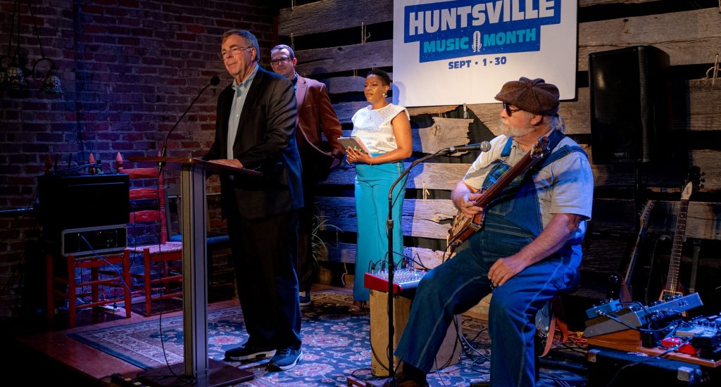 Huntsville Mayor Tommy Battle delivers remarks at the Huntsville Music Month announcement at Tangled String Studios. Joining him are Music Officer Matt Mandrella, Huntsville artist Karmessa Padgett and Microwave Dave, who is holding a cigar box guitar. There is a large sign at the back of the stage that says Huntsville Music Month.