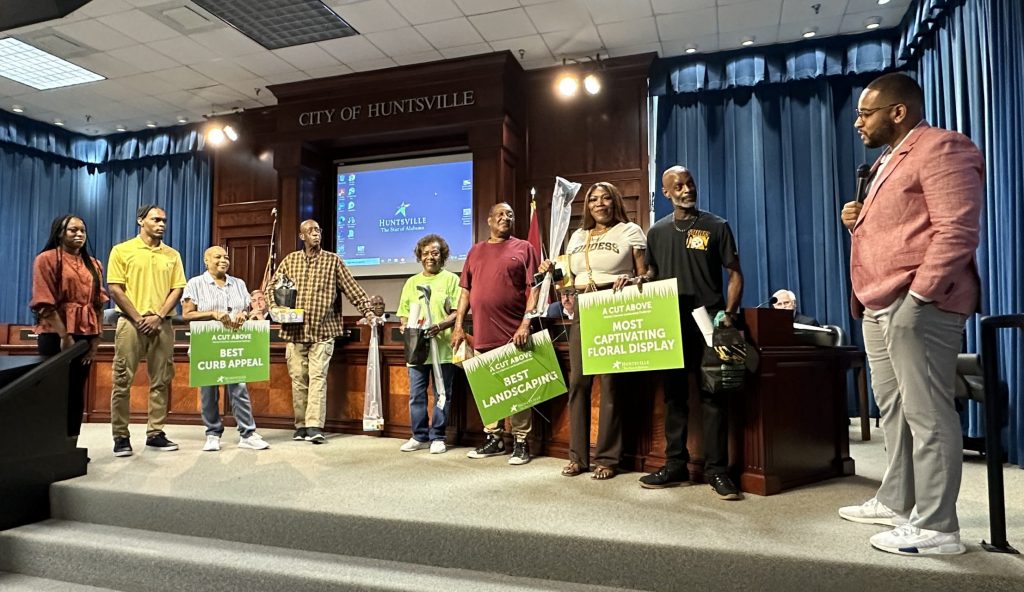 a group of men and women standing on the dais in a meeting room holding green yard signs and prizes they received from a man holding a microphone