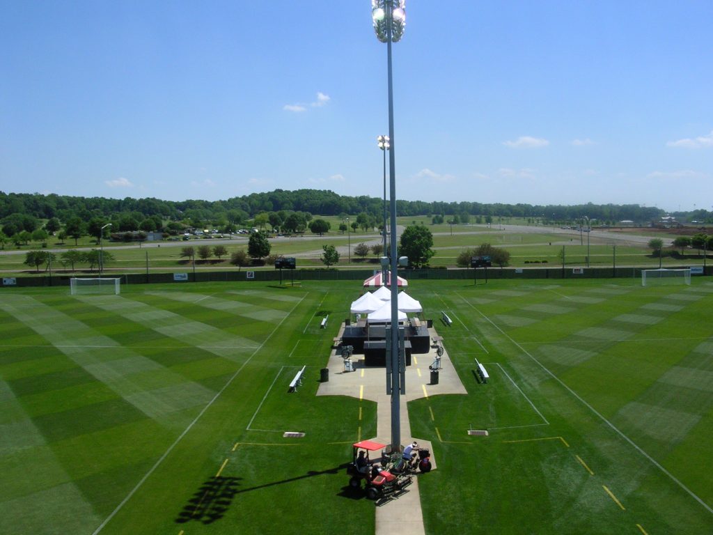 green turf soccer fields separated by stadium lights, press box and team benches