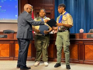 Man in blazer presents proclamations to two teenage Eagle Scouts at City Council meeting