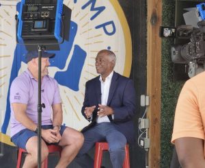 Man in blue blazer speaks to man with purple collared shirt with cameras and THE CAMP mural in background