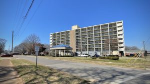 A large apartment complex is seen next to a road. There is blue sky overhead and power lines can be seen.