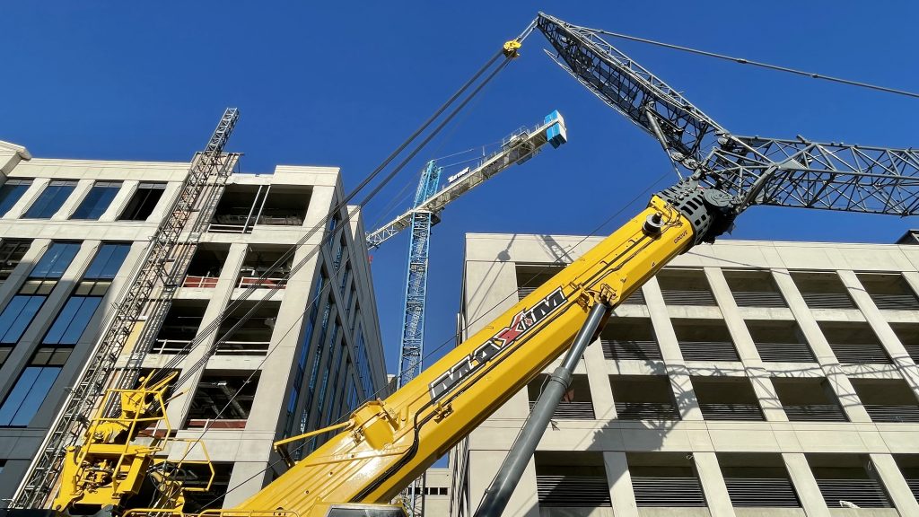 A large crane disassembles a tower crane used in the construction of the new Huntsville City Hall on Fountain Circle. The crane in the foreground is yellow, while the tower crane is blue and white. There are large buildings on either sides of the cranes and blue sky overhead.
