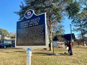 A historic marker gives the history of the Edmonton Heights neighborhood. There is a tree in the background and two people talking.