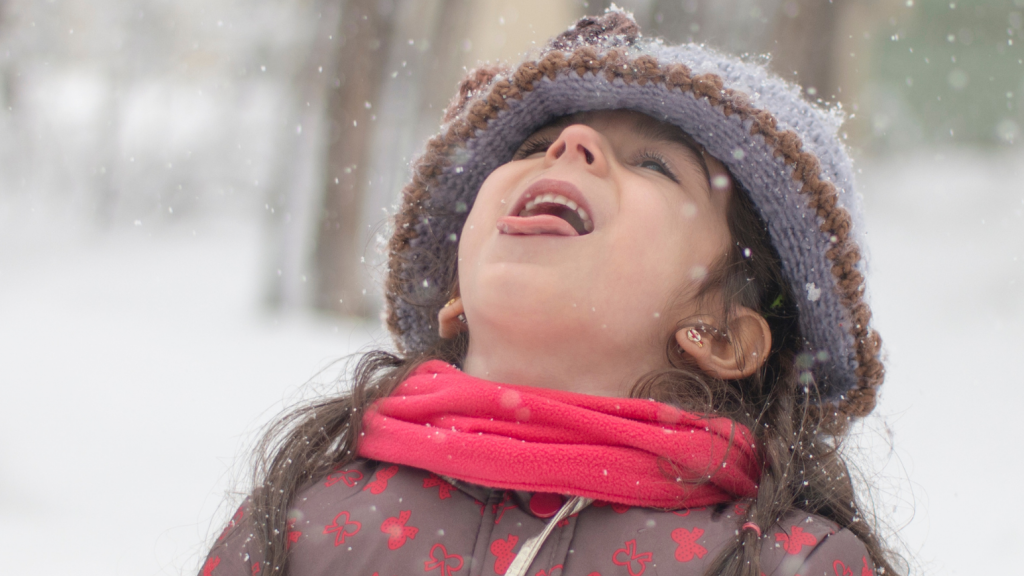 A young girl catches snowflakes on her tongue. She's wearing a wool cap and scarf.