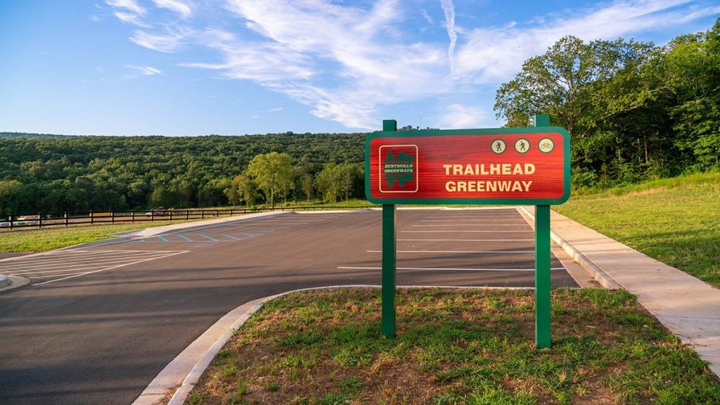 A picture of the Trailhead Greenway sign with mountains and a parking lot in the background.