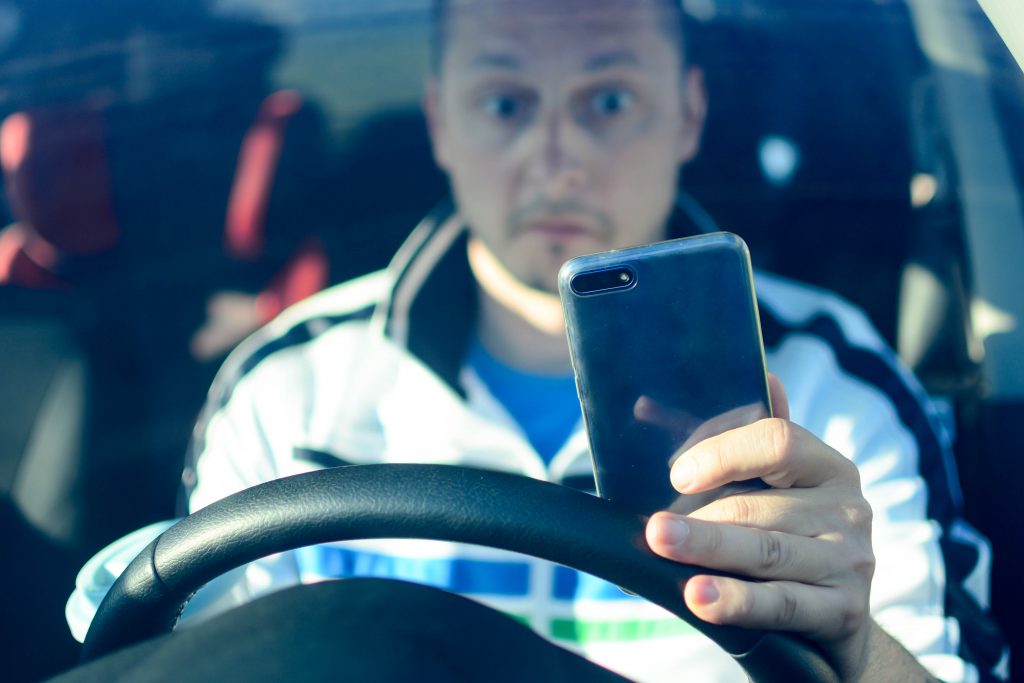 A man looks at his phone while driving. He has a surprised look on his face.