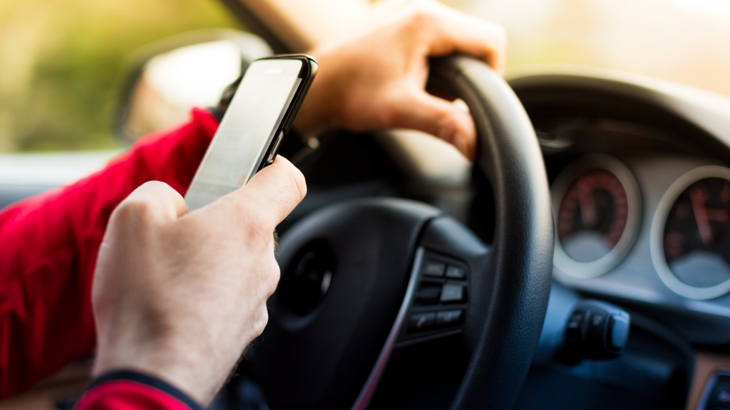 A white person holds a phone while steering a vehicle. The person is wearing a red shirt. The phone is black.