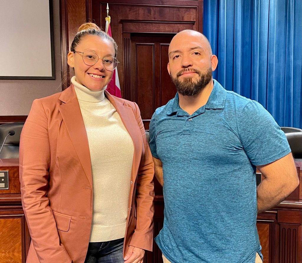 a woman in a tan jacket and white turtleneck with her hair pulled back stands next to a man wearing a blue shirt in front of a desk in CityHall