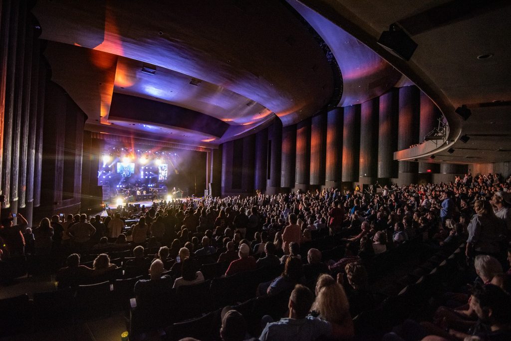 Attendees watch a concert at the Mark C. Smith Concert Hall at the Von Braun Center. There are colored lights reflecting off the walls.