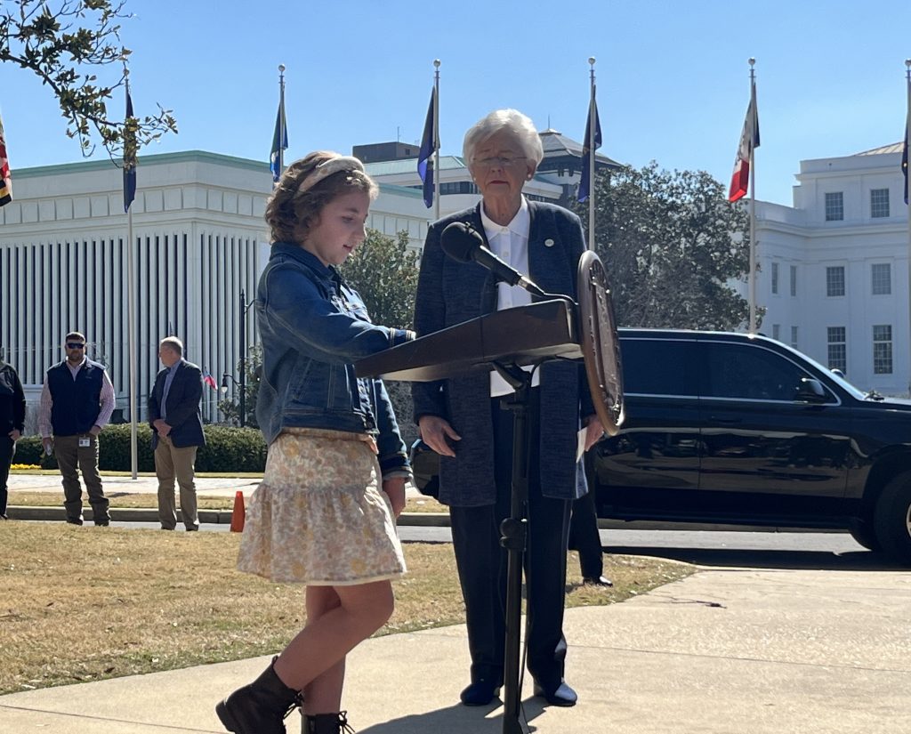 girl wearing a skirt and blue jean jacket stands next to an older lady wearing a blue suit in front of the alabama state capitol building. the girl is standing by a microphone giving a speech