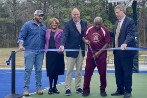 James Crawford cuts the ribbon on the newly renovated James C. Crawford Park. He's wearing a red athletic suit with an Alabama A&M logo. He's joined by other people, who are wearing suits. They are holding a blue ribbon.