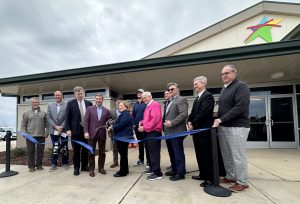 line of people behind a blue ribbon. a person with large scissors is cutting the ribbon, in front of a building