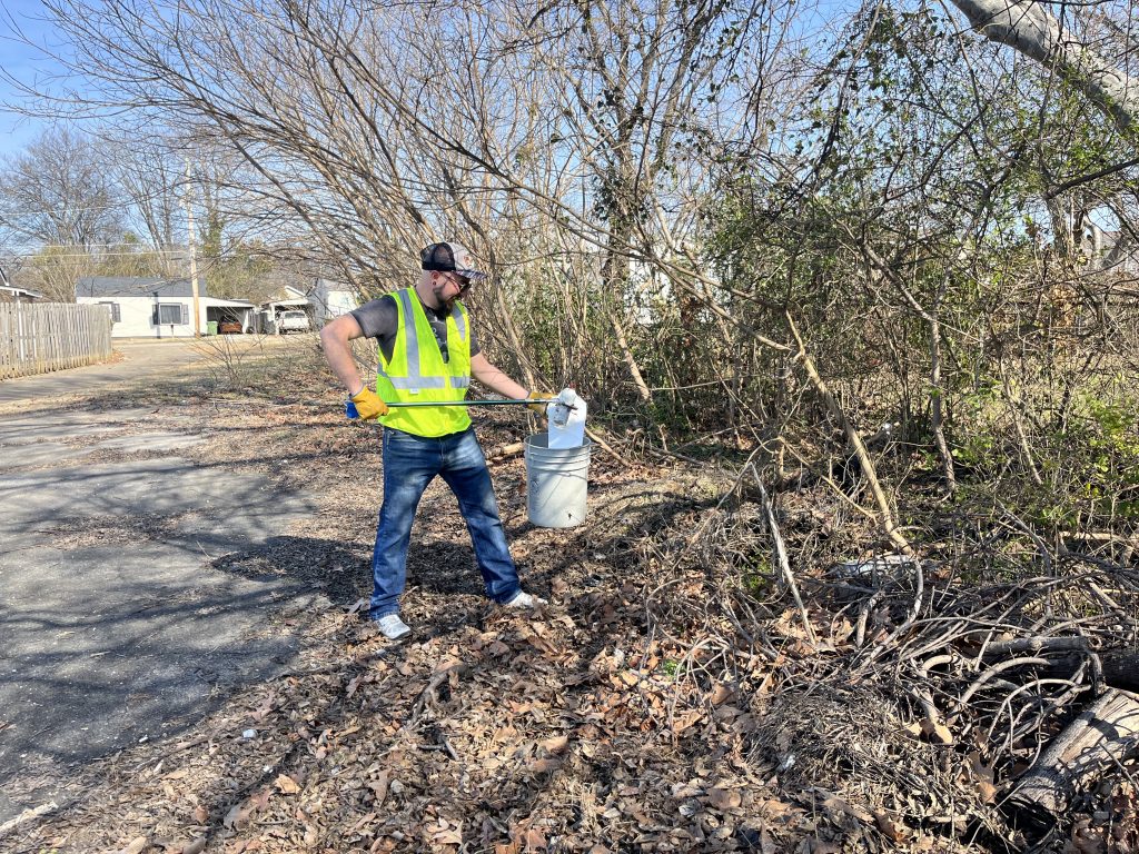 man wearing blue jeans and a neon yellow vest uses a stick and garbage sack to pick up litter from the side of the road