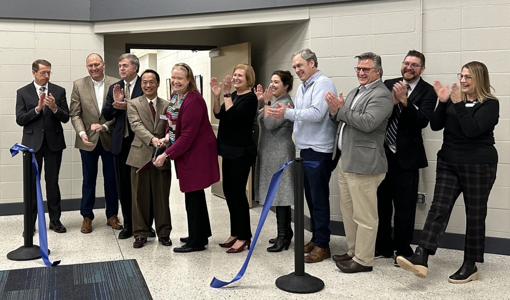 a group of men and women standing in line in a room. One is holding a large pair of scissors, having just cut a long blue ribbon which is floating to the floor. everyone is clapping