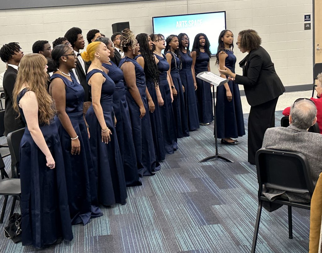 teenage men in tuxedos and women in long blue gowns stand in front of a conductor to sing a song in the new rehearsal room