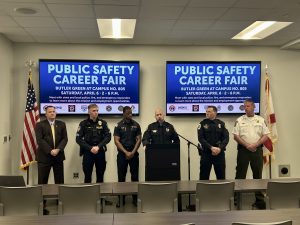 Six law enforcement officers standing with a Huntsville Police Department officer standing at a podium to announce the Madison County Public Safety Career Fair at the Huntsville-Madison County 911 Center