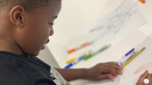 A child works on art using markers, which are near his hands.