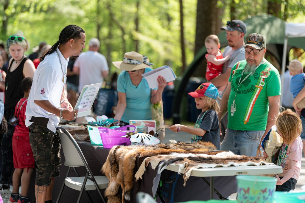 Earth Day attendees visit a vendor's table at the 2022 Earth Day event at Hays Nature Preserve. The vendor at left is wearing a white shirt and camouflage pants. There are parents and children to the right. There appears to be animal skins on the table.